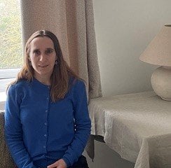 Toni Hoskins is sitting, smiling. Toni is a white woman with light brown hair and is wearing a blue cardigan. There is a table and lamp in the background.