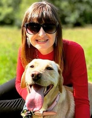Fern, (a white woman in her thirties with long brown hair) sits in a green field with her golden, Labrador guide dog, Nancy. Fern wears a bright red top and sunglasses and is smiling towards the camera. Nancy has her pink tongue sticking out happily, as she lays in the grass with Fern.