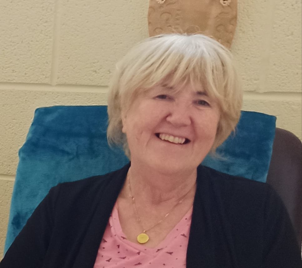 A lady with blonde short hair is smiling, sitting on a chair with a cream concrete wall behind her. She's wearing a pink top, navy blue cardigan, and has a gold necklace on.