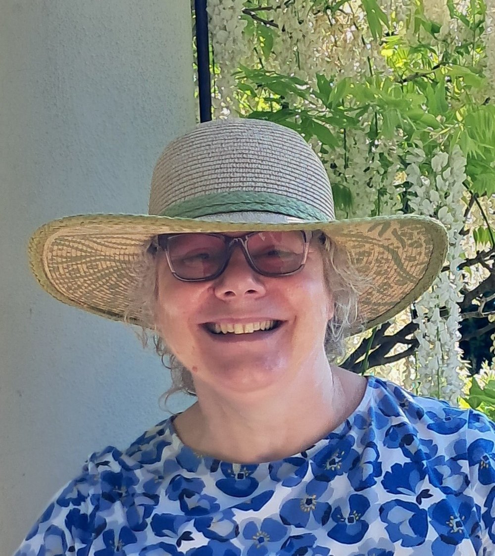Head shot of Delia wearing a straw hat.  
Wisps of light hair peeping out from under a straw hat. 
She is wearing light pink square framed glasses with tinted lens. 
Smile showing teeth and being happy. 
White wisteria plant in the background