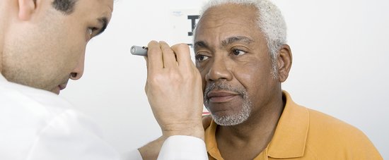 A man having an eye test. He has a focused expression, grey hair and a grey beard and is wearing a pale orange coloured polo shirt. An optician, standing to the left, shines a light from a small torch into his eyes. The optician is looking at the man’s eyes. Behind them is a plain white wall.