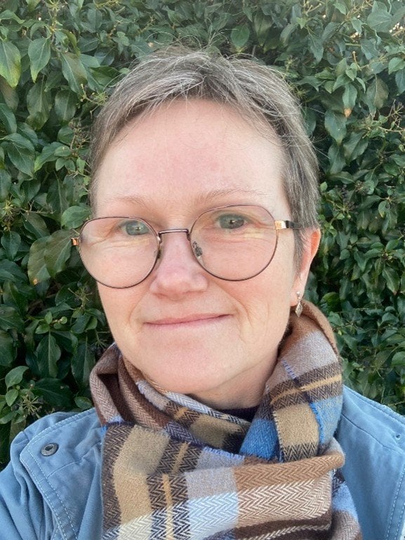 Ruth Williams, a woman with short grey hair and glasses, is smiling in front of a foliage wearing a light blue jacket and tartan scarf.