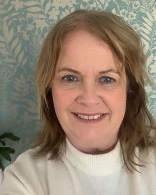 A woman with shoulder-length dark blonde hair and blue eyes is smiling. She's wearing a white high neck jumper and standing in front of a floral patterned wallpaper.