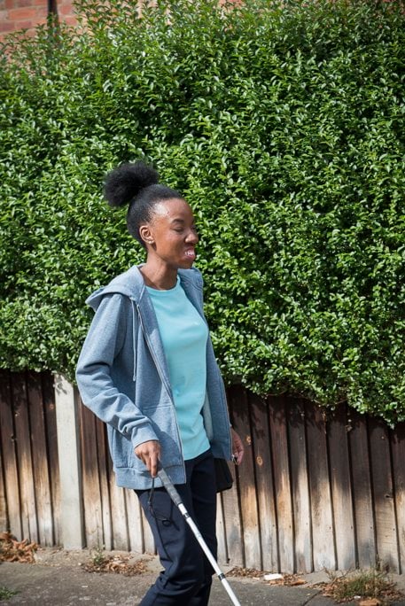 Blind psychotherapeutic counsellor, Sandra, walking along wooden fence with her white cane wearing a blue t-shirt and a grey zip-up hoodie.