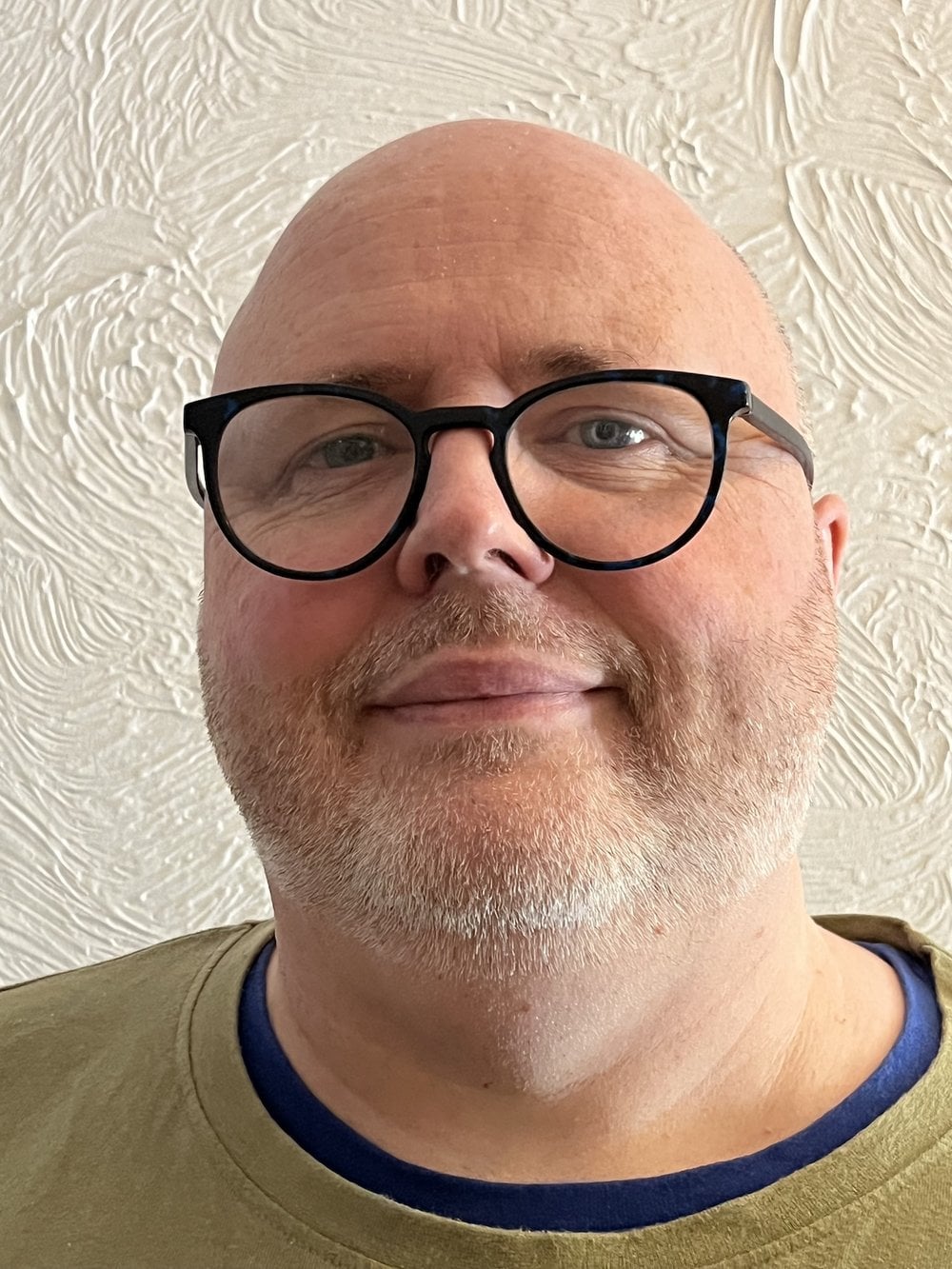 Stood in front of a white woodchip wall. Ken is warm and welcoming, you can see his big smile and has 'smiley eyes' behind his blue rimmed glasses, his full round face is bearded, and you can see his bald head, you can see his green v neck jumper with a dark t-shirt popping through the neckline