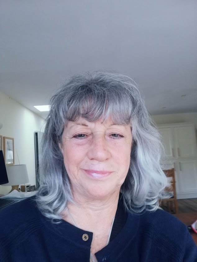 A close-up portrait of a woman with shoulder-length grey hair and wearing navy blue, looking into the camera with a gentle smile, indoors in natural light.