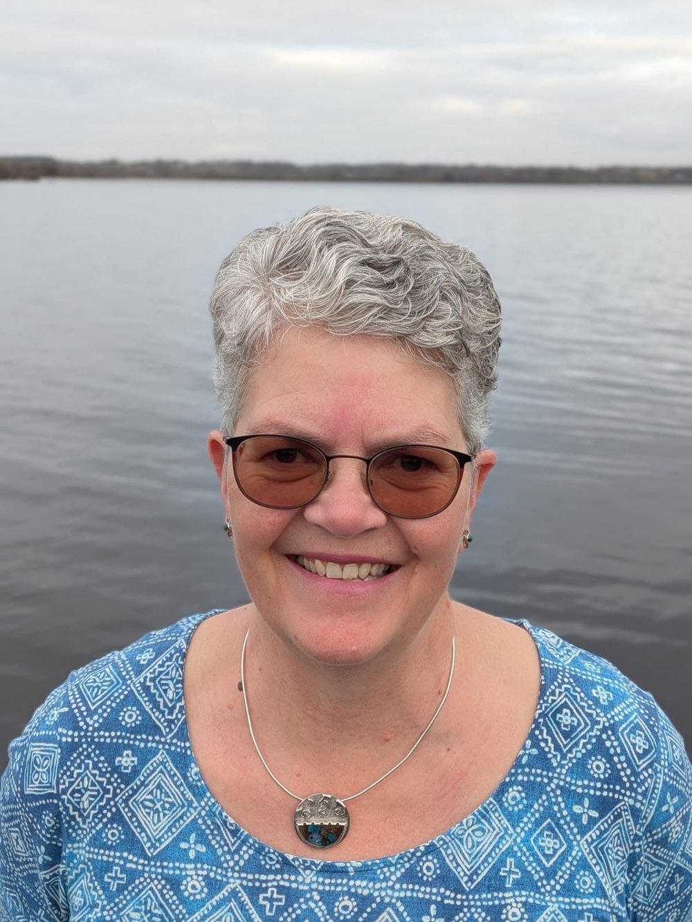 Sonja Keaney with grey wavy hair smiling at the camera. Wearing tinted glasses and a green top, there is water in the background