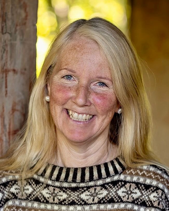 A professional outdoor head-and-shoulders photograph of Margot smiling, with long blonde hair, wearing a Fair Isle patterned jumper. Trees are visible in the softly blurred background.