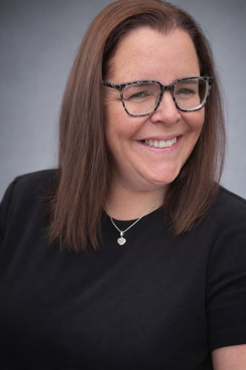 Photo is of Clare and she has medium brown shoulder length hair and black rimmed glasses. She is smiling. She is wearing a love heart pendant in a silver chain. She is wearing a black top.
