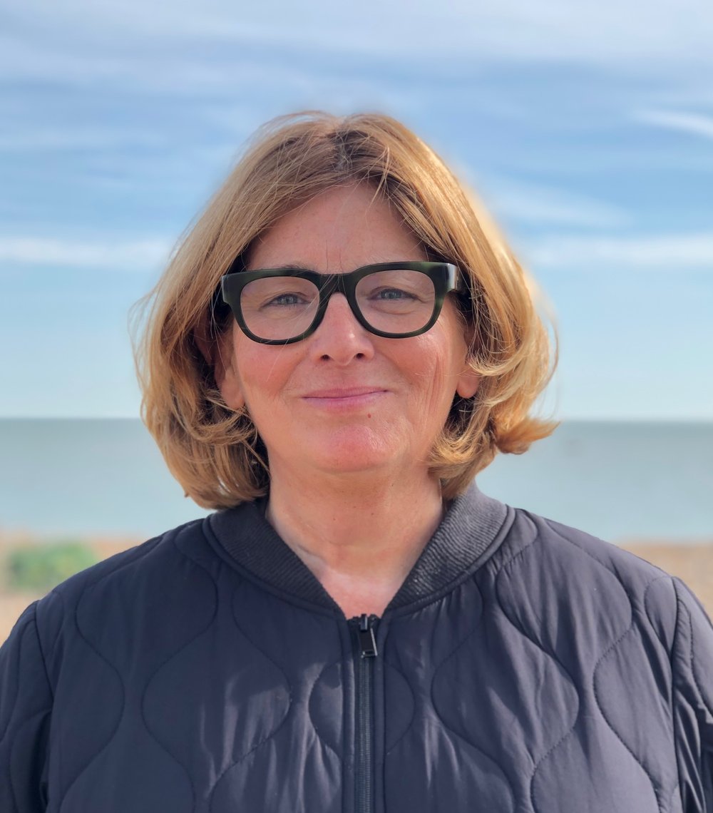 Half-length photo of a woman facing the camera, she has brown shoulder length hair, is smiling, wearing black glasses and a black padded jacket. The sea is in the background of the photo.