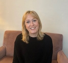 Headshot of Lucy Wykes, a RNIB Accredited Sight Loss counsellor. Lucy is a white woman with blonde mid-length hair smiling warmly. She is wearing a black jumper and sitting on a red armchair.