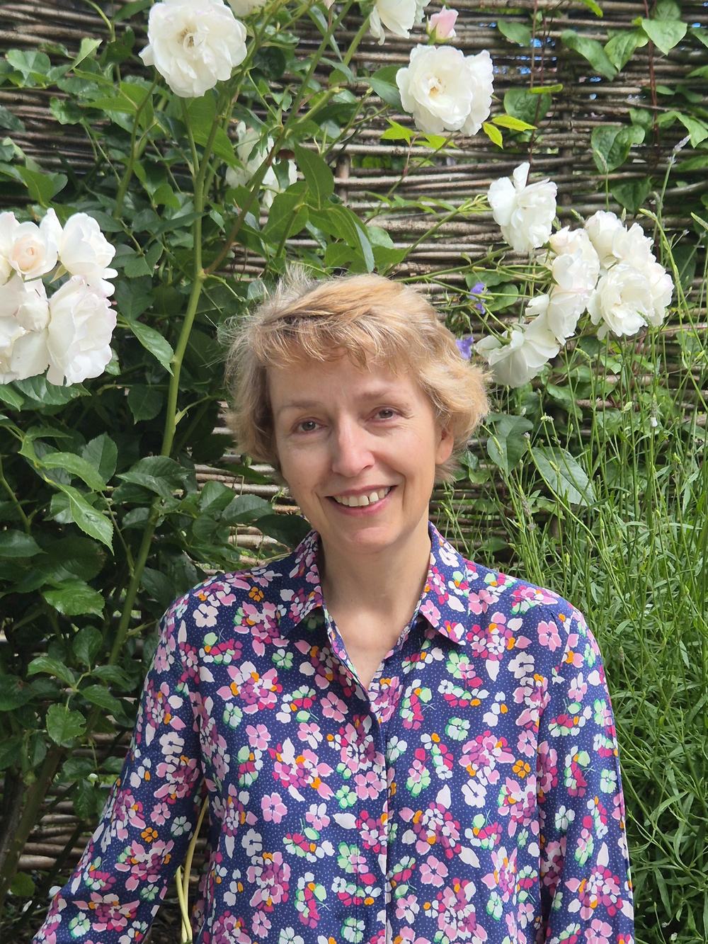 An image of Janet Reed smiling, facing the camera. There is greenery and flowers behind Janet. She is wearing a purple shirt with a multi-coloured floral print, and has light short hair.