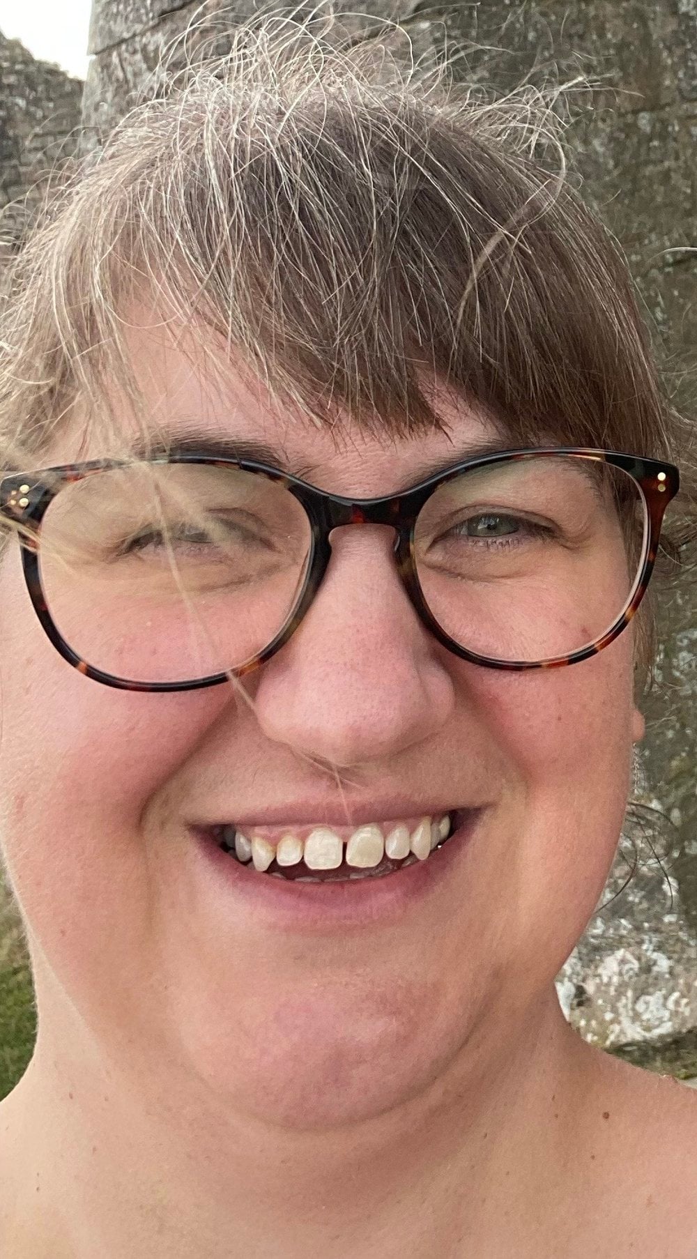 A close up photograph of Paula taken outdoors with her smiling at the camera, she has light brown hair with bangs across her forehead and she is wearing dark round framed glasses. A strand of hair falls gently across one lens. She has a natural smile. The background is a stone wall giving the image a relaxed setting.