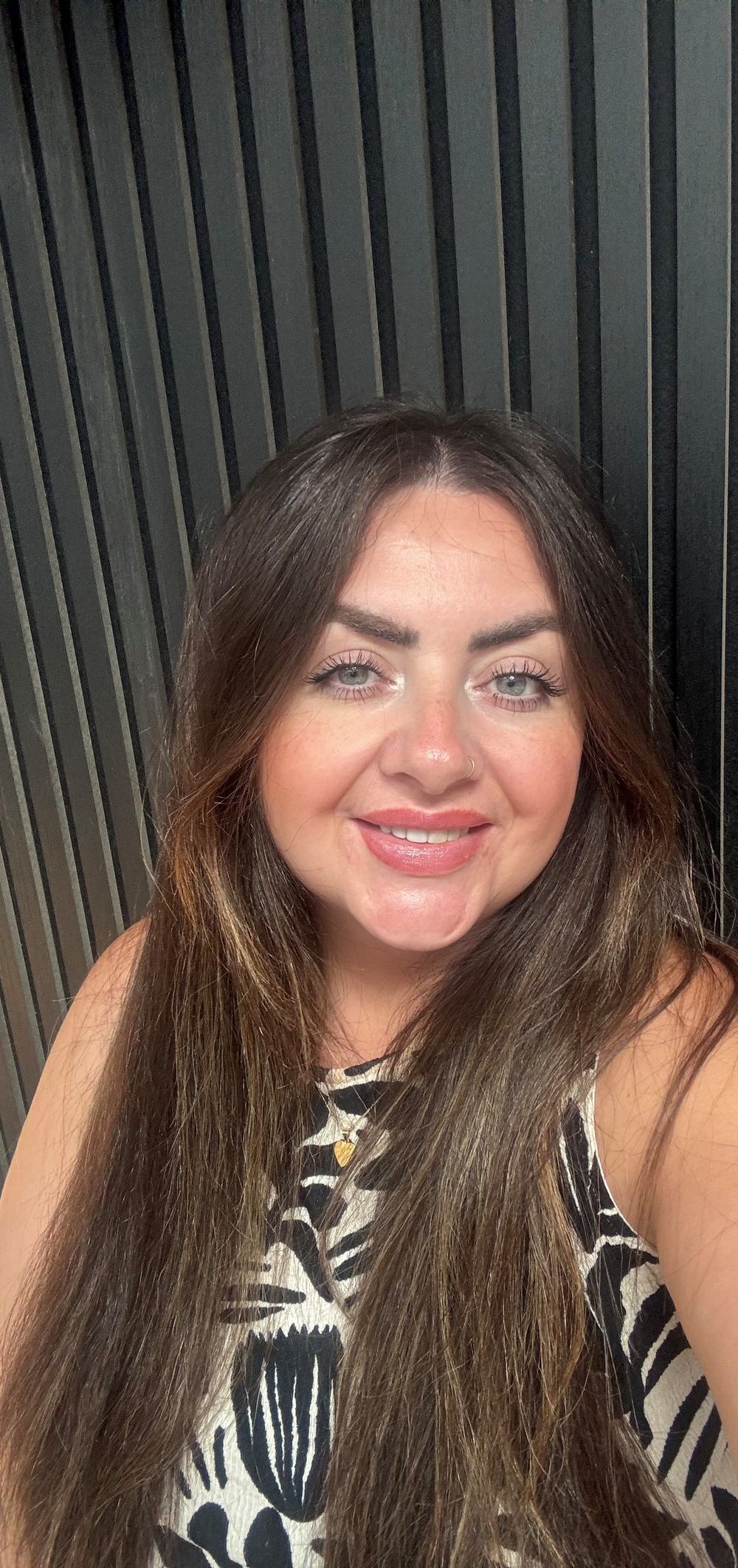 The image is of Kate smiling to the camera in front of a black background, they have long dark hair, fair skin and green/grey eyes. They am wearing a beige and black top.