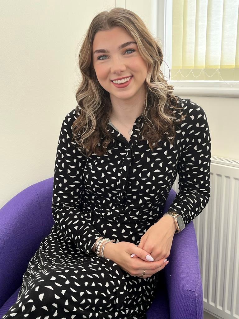 A smiling woman with shoulder-length wavy brown hair featuring blonde highlights sits in an armchair in a bright counselling room. She is wearing a long-sleeved black dress with a white abstract pattern, a silver smartwatch, and stacked bracelets. Her hands are gently clasped in her lap, and she is looking directly at the camera with a warm, welcoming expression. A window with cream blinds and a white radiator are visible in the background.