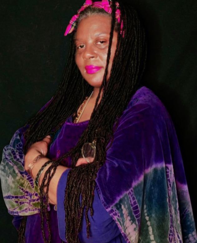 Portrait photograph of a woman with long black dreadlocks, pink headband and lipstick, wearing a purple velvet outfit against a dark background. Crossed arms, layered bracelets, patterned sleeves, and confident expression.