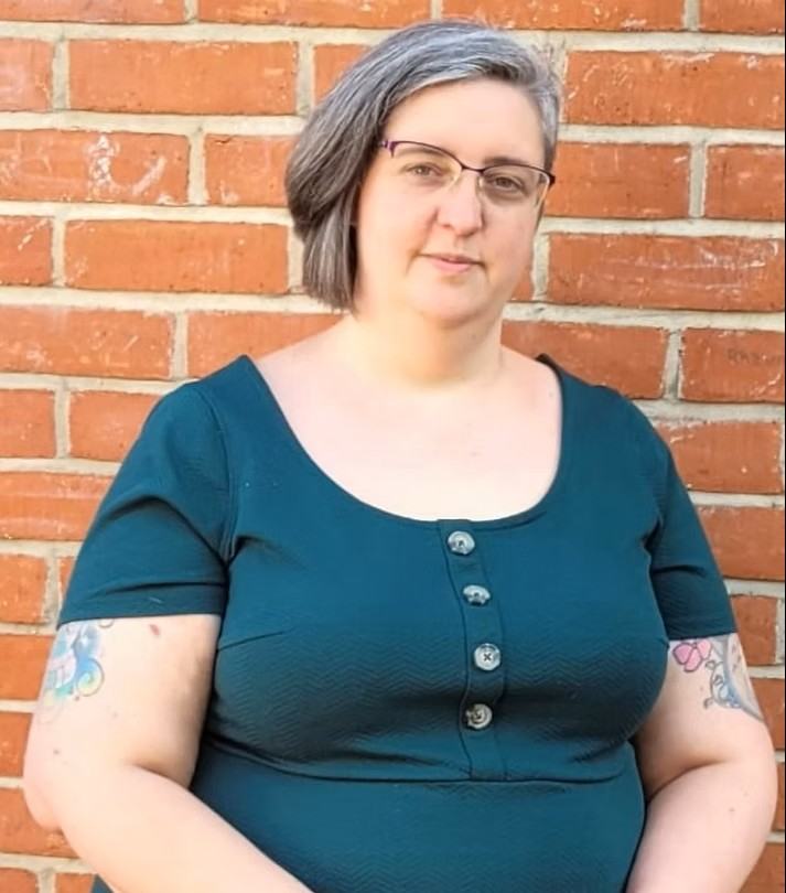 Jenni - a plus-sized white woman, with short brown hair and glasses. She is wearing a dark green dress. She has tattoos on her arms. She is stood in front of a brick wall.