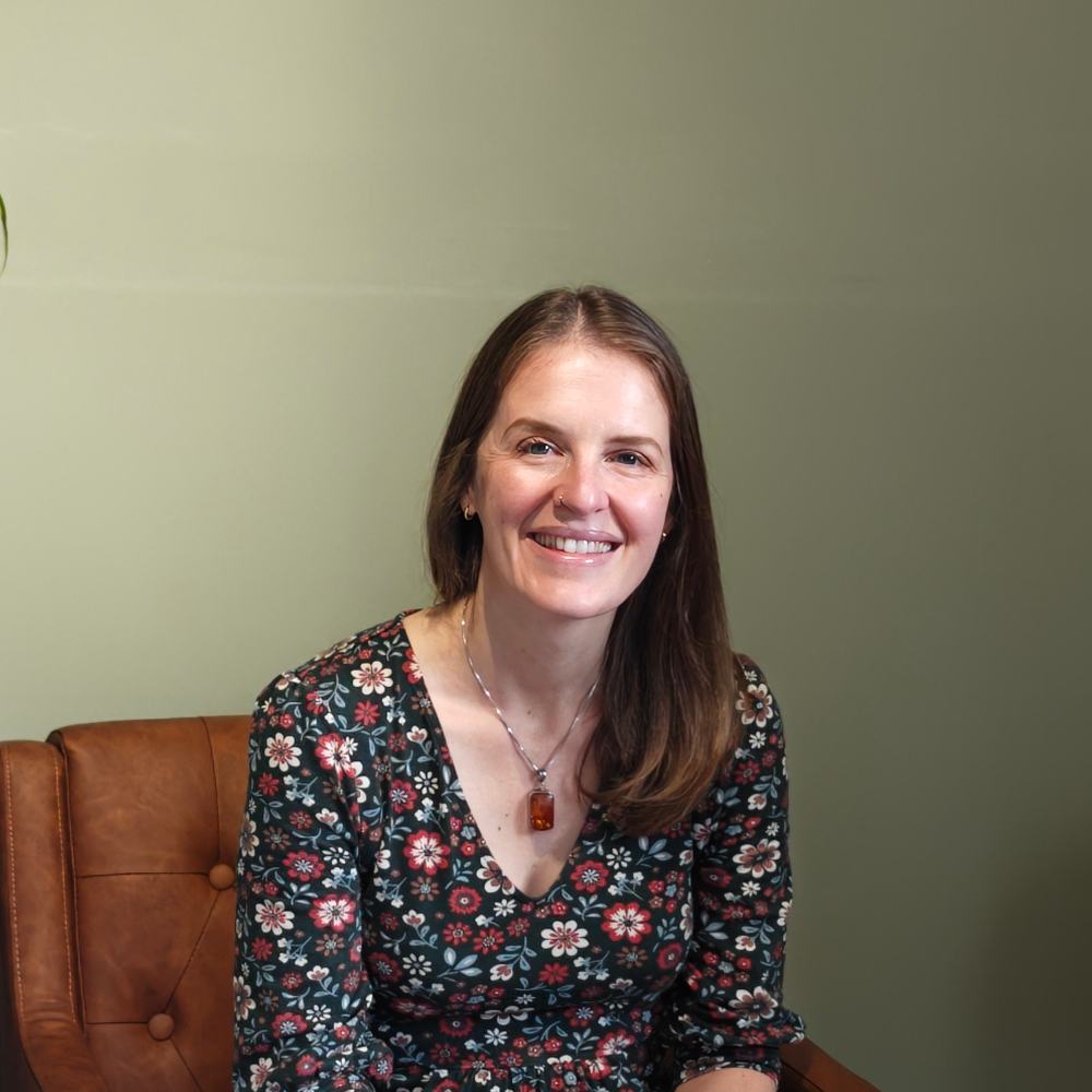 Head and shoulders photo portrait of Claire Bell - I have long brown hair with a side parting swept over one shoulder. I am smiling. I am wearing a green dress with a pattern made up of yellow and white flowers and a silver necklace with a rectangular amber pendant. I am sitting on a brown leather chair and the wall behind me is painted green.