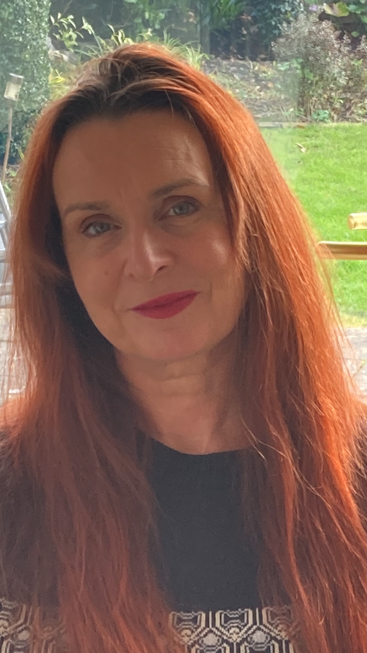 An image of Deborah McDonnell, smiling, facing the camera. In the background is a garden. Deborah has long red hair, is wearing a black jumper, and pink lipstick.