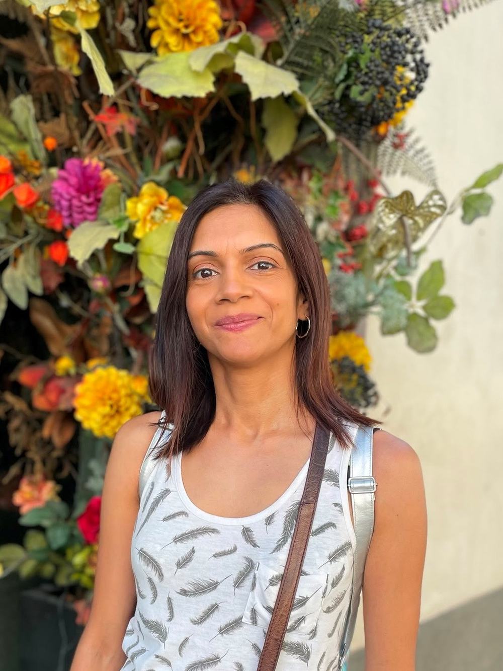 Woman in her forties, dark hair, smiling, standing in front of a display of colourful flowers.