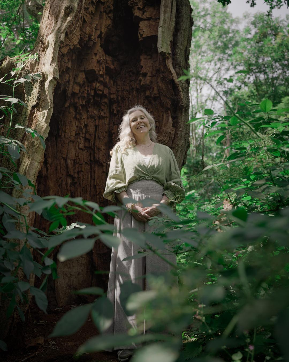 An image of Helen Curzon standing in a forest. There is a large tree behind her and she's smiling at the camera. Helen has light long hair, a green top and cream trousers, with her arms clasped in front of her.