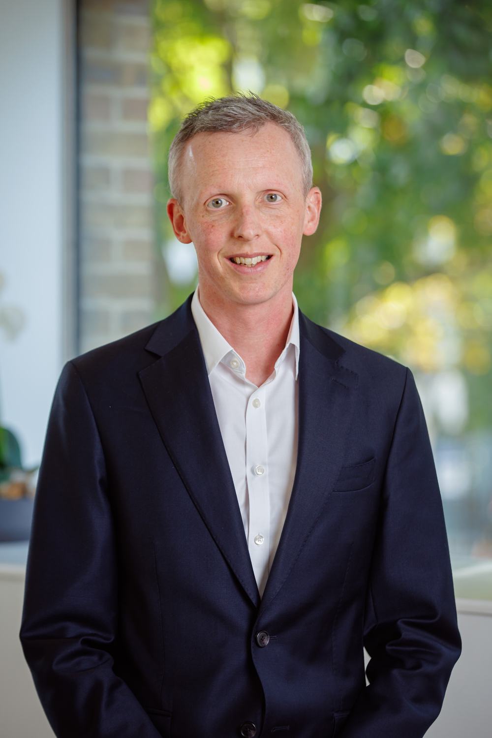 A picture of Andrew McCracken, a man with grey hair, wearing a navy suit with white shirt, smiling at the camera.