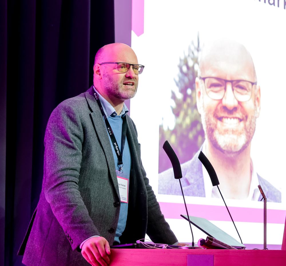 A picture of Nigel Fletcher standing giving a speech at a lectern, he is a bald man with glasses and a beard, wearing a grey blazer and light blue jumper and shirt. Behind him is a presentation with a picture of him.