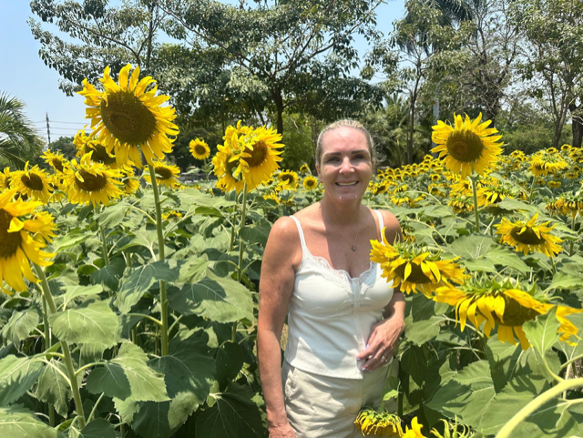 Cindy in a sunflower field on a sunny day. She's wearing a white camisole and her hair is tied back.