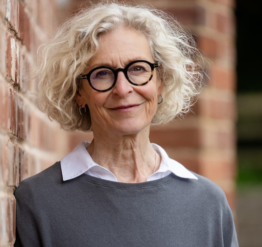 An image of Caroline Midmore, facing the camera smiling. Caroline has short light hair and is wearing a white shirt and grey jumper, small earrings, and black glasses.