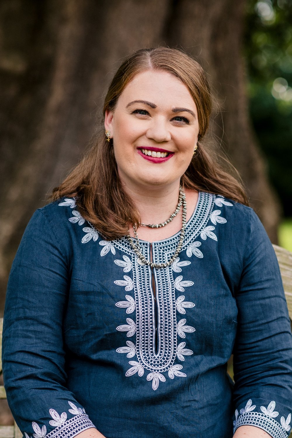 A head shot of Emily, she is sitting under a tree and is wearing a blue top with white detail. She is smiling.