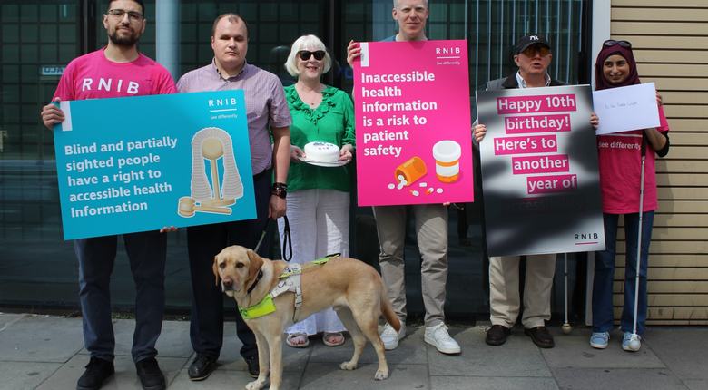 A group of six RNIB supporters stand outside the Department of Health and Social Care with placards to mark the tenth birthday of the Accessible Information Standard. One sign reads, “Blind and partially sighted people have a right to accessible health information,” while another states, “Inaccessible health information is a risk to patient safety.” A guide dog stands in the foreground. Another placard celebrates “Happy 10th birthday, here's to another year of" with the remaining words blacked out to show the impact of diabetic retinopathy on reading information.