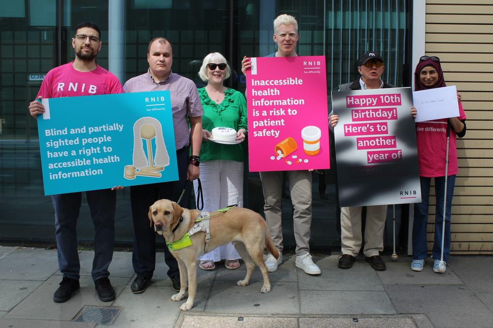 A group of six RNIB supporters stand outside the Department of Health and Social Care with placards to mark the tenth birthday of the Accessible Information Standard. One sign reads, “Blind and partially sighted people have a right to accessible health information,” while another states, “Inaccessible health information is a risk to patient safety.” A guide dog stands in the foreground. Another placard celebrates “Happy 10th birthday, here's to another year of" with the remaining words blacked out to show the impact of diabetic retinopathy on reading information.