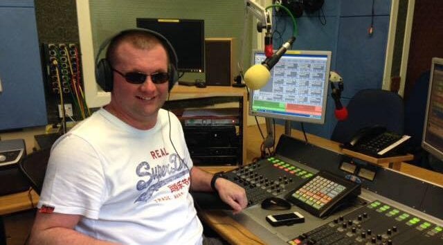 Alan MacDonald sits at a radio desk, smiling. He is wearing headphones and dark glasses.