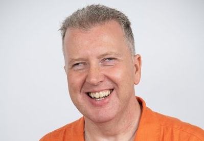 Close up of Allan infront of a white background, wearing an orange top.