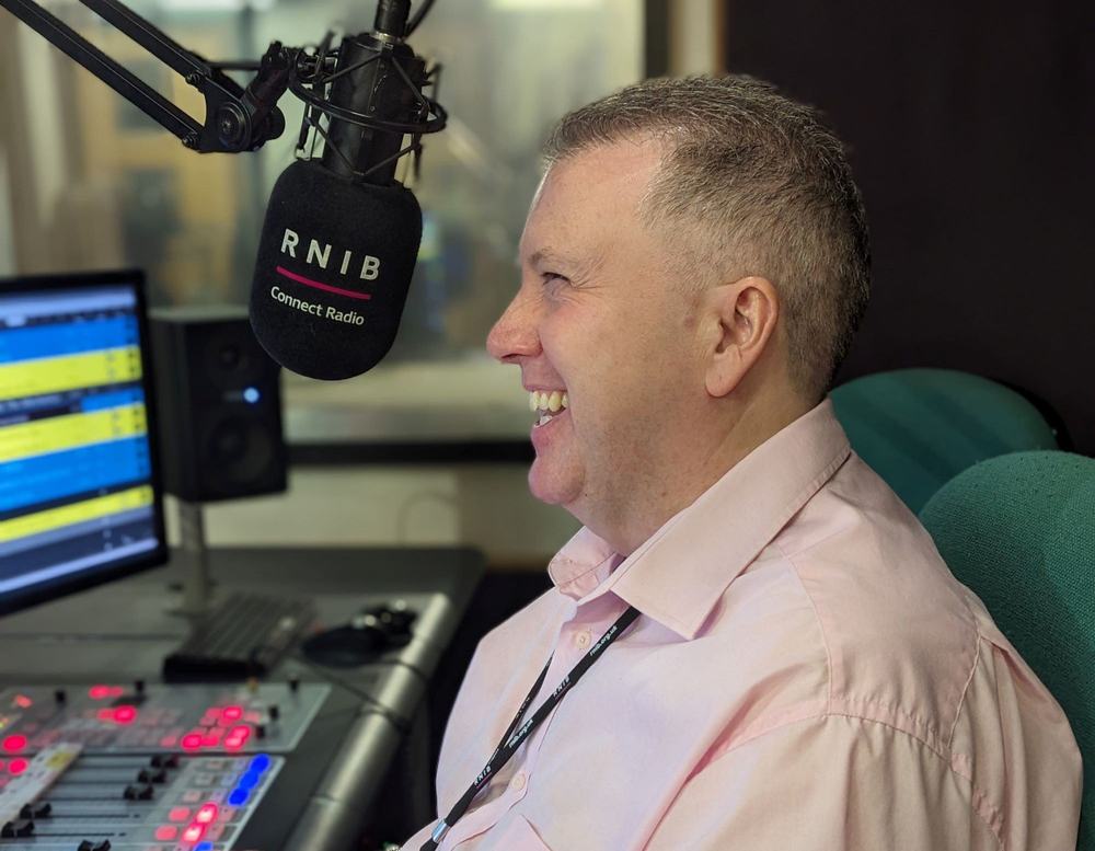 Allan in the studio laughing as he gets set up to present. He's wearing a dashing light pink short sleeve shirt.