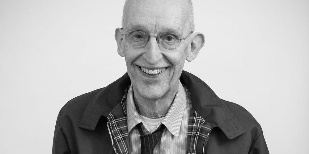 A black and white photo of Aly smiling. He's slim, wearing rimless glasses, a loose tie over a dress shirt and a check lined jacket over the top.