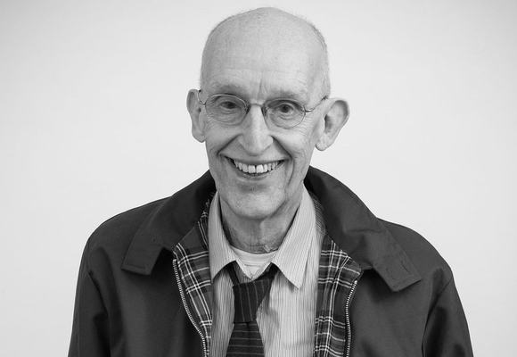 A black and white photo of Aly smiling. He's slim, wearing rimless glasses, a loose tie over a dress shirt and a check lined jacket over the top.