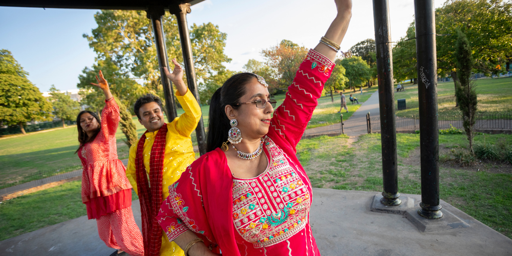 A woman is in the centre of the image in a red colourful dress dancing with her left arm raised. There's a man and woman also dancing behind her. They are in a park, with grass, trees, and clear skies in the background.