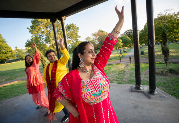 A woman is in the centre of the image in a red colourful dress dancing with her left arm raised. There's a man and woman also dancing behind her. They are in a park, with grass, trees, and clear skies in the background.