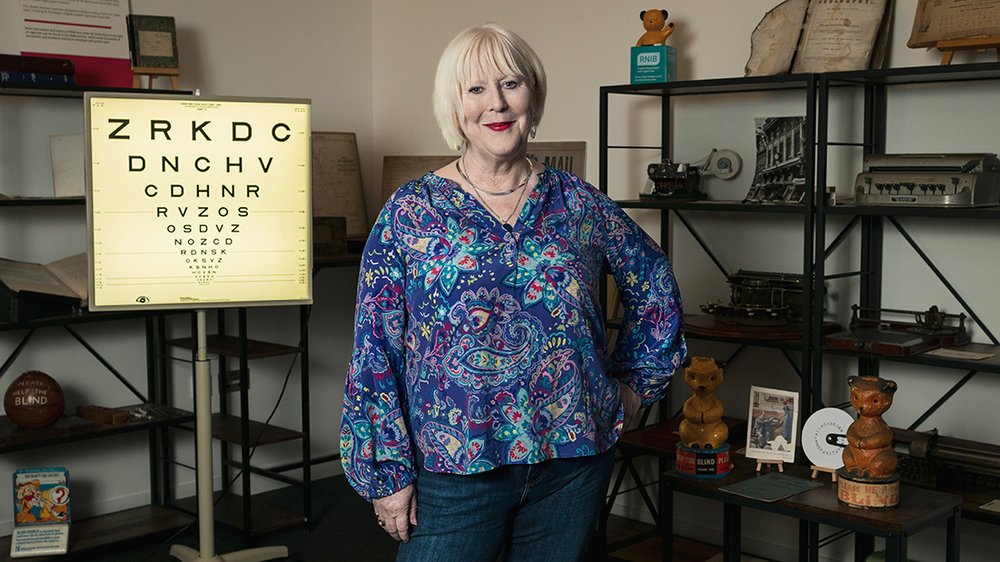 This portrait is of Anna, a middle-aged woman with fair skin and blonde hair, standing confidently in RNIB’s heritage space dressed in a vibrant blue and purple paisley blouse and jeans, with a gentle smile and a relaxed, welcoming posture. Her blonde bob haircut and red lipstick add a classic touch to her style. To her right, there’s a vintage illuminated eye chart featuring large letters at the top and smaller ones toward the bottom. The background is filled with shelves displaying a variety of RNIB artefacts and nostalgic objects – including vintage Sooty coin boxes, books, framed photographs and curiosities. The setting feels personal, curated, educational and archival. The overall atmosphere is one of quiet confidence, charm, and curiosity – a hint of Anna’s rich personal and professional story.