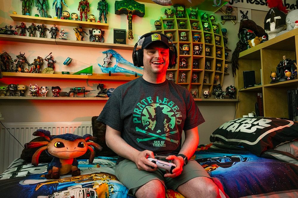 This portrait is of Ben, a man in his early thirties with fair skin, is seated comfortably on a Star Wars-themed bedspread in his bedroom filled with collectibles and gaming memorabilia. He is wearing a dark grey “Pirate Legend – Sea of Thieves” t-shirt, khaki shorts, a baseball cap, and a large gaming headset. His baseball cap features the SightlessKombat logo patch – this is the handle he works under in gaming, and the logo is a graphic of an eye with two swords crossed in front of it. He holds a game controller in both hands and is smiling broadly, exuding happiness and confidence. The lighting in the room includes red and green hues that cast a dynamic glow across the scene, enhancing the atmosphere.
Behind him, the room is richly decorated with shelves full of action figures, gaming props, and vinyl figures. A plush toy of Nix, a character from the video game Star Wars Outlaws, sits at the edge of the bed next to Ben. Overall, this joyful photo captures Ben’s enthusiasm for gaming culture, his immersive setup and his individuality.