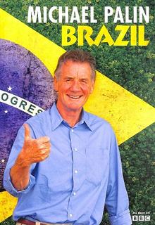 Book cover for Michael Palin: Brazil. A smiling older man stands in front of a large Brazilian flag backdrop, giving a thumbs-up gesture. He wears a light blue button-up shirt with sleeves rolled up. The title appears at the top in large white and yellow letters, with “MICHAEL PALIN” above “BRAZIL.” Behind the flag, a lush green forest is visible. The overall design uses the colours of the Brazilian flag—green, yellow, blue, and white—and conveys a positive, travel-themed atmosphere.
