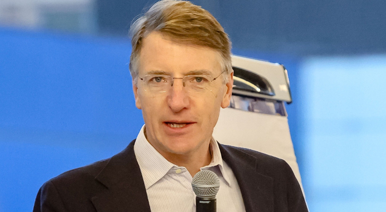 Sir Charlie is wearing a smart black jacket and trousers and a white shirt with the top button undone. He is a middle-aged intelligent looking man with glasses and mousy brown/greying hair. He is holding a microphone as if in full speech mode.