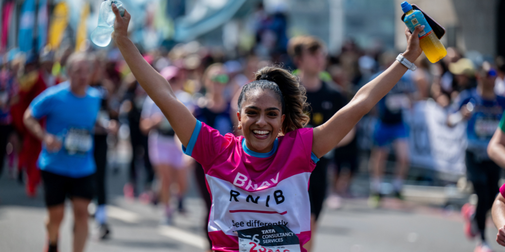 A person smiling with their arms in the air holding water bottles, taking part in a Challenge Event wearing an RNIB t-shirt.