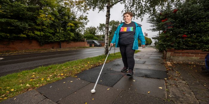 Charmaine a white woman with short brown hair wearing a bright blue waterproof jacket and a black t shirt with coloured pencils on it walking on a residential street with a cane