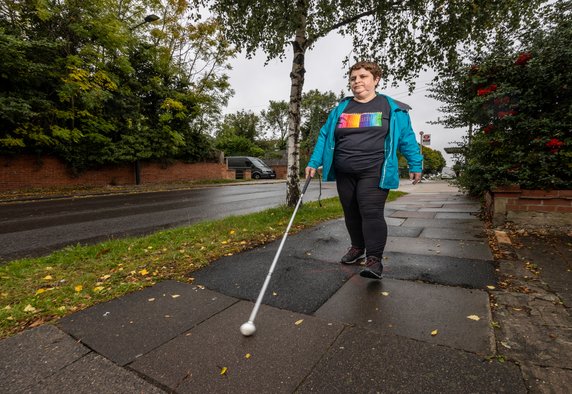 Charmaine a white woman with short brown hair wearing a bright blue waterproof jacket and a black t shirt with coloured pencils on it walking on a residential street with a cane