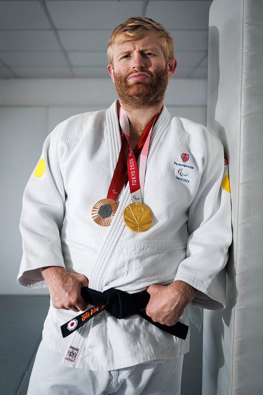 Chris, a man in his early thirties with fair skin, short strawberry blond hair and a darker blond beard, in a powerful and focused portrait proudly wearing his judo uniform and two Paralympic medals around his neck — a gold from Tokyo 2020 and a bronze from Paris 2024. His confident, intense expression, along with his athletic stance and build, convey strength, determination, and hard-earned accomplishment.
Chris is leaning casually against a padded wall, hands gripping the edge of his black belt — a symbol of mastery and dedication in martial arts. His judo uniform bears patches for Paralympics GB and Tokyo 2020, highlighting his national representation and elite level of competition.
He stands in a martial arts training centre, with clean white walls and soft lighting that brings all attention to Chris himself. His blond hair and beard are slightly tousled, and there’s a visible sense of calm control in his posture — the kind of quiet confidence that comes from years of discipline and performance under pressure.