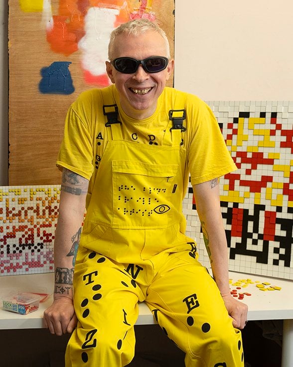 This portrait is of Clarke, a man in his mid-forties with fair skin and blond hair, sitting casually on the edge of a table in his studio. He’s dressed head-to-toe in bright yellow – a matching t-shirt and overalls ensemble – adorned with black graphic elements including braille dots, letters, and symbols. Clarke wears dark sunglasses and has visible tattoos on both arms, adding to his distinctive and expressive look. His wide, genuine smile radiates warmth and approachability. Behind Clarke are his artworks – colourful, pixel-art-style mosaic panels – made from small multi-coloured, square tiles. A partially painted board sits propped up behind him. On the table beside Clarke sits a container of LEGO-style pieces, further reinforcing the creative, tactile, and accessible nature of Clarke’s art. This portrait feels vibrant, joyful, dynamic, and rooted in personal expression –  a celebration of individuality and art as a language beyond the visual.