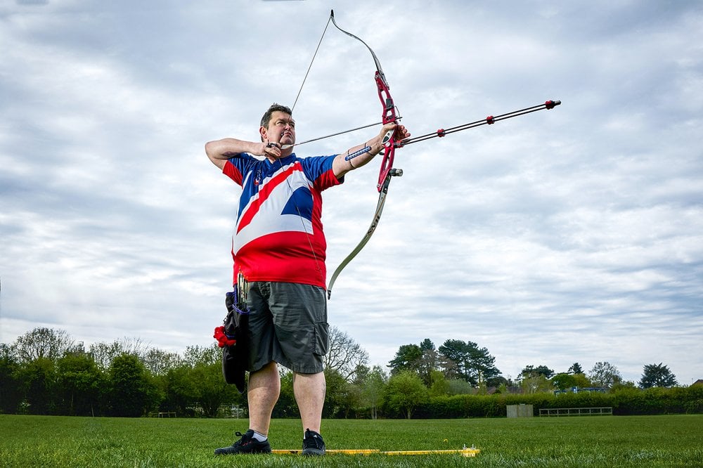 This portrait is of Clive, a man in his late forties with fair skin and short dark hair, outdoors, standing on a grassy field under a dramatic cloudy sky, engaged in the sport of archery. He is in the middle of drawing his bow, with the arrow pulled back, ready to release.
He wears a red, white, and blue shirt featuring a large graphic design which is part of the Union Jack. He also wears shorts and has equipment attached to his belt, including arrows and accessories for archery.
His expression is focused and determined, and his posture demonstrates skill and discipline, highlighting his commitment to the sport. The surrounding environment, with trees in the background and open space, creates a fitting setting for his target practice.