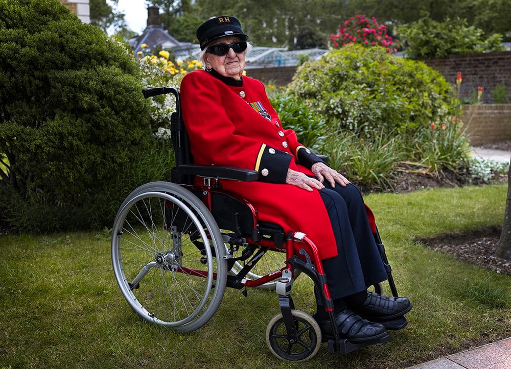Connie, a 102 year old woman with fair skin,  sitting in a wheelchair, wearing the iconic bright red coat and navy-blue uniform of the Chelsea Pensioners – a retirement and nursing home for British Army veterans. Connie, who is adorned with medals on her chest and dressed immaculately, exudes pride and composure. She is wearing dark sunglasses and a black cap embroidered with the initials "RH" (Royal Hospital). Her hands rest gently on her lap, and her posture is relaxed yet noble.
The setting is a well-tended garden, with flowers and lush greenery creating a beautiful background. The image captures a moment of peaceful reflection, reverence, and timeless service, honouring the life and legacy of someone who has served her country with distinction.